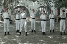 Seven people dressed in complete white outfits with thick black belts across their midriffs and tiny red hats perform a dance or performance