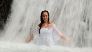 A woman in a white dress stands at the base of a waterfall with her arms outstretched as if to let the water cleanse her