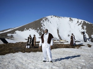 In this still of Christopher Ulutupu's video work the artist's father stands on a sunny, snowy mountain peak wearing a dapper white suit, sunglasses and fur throw, looking up towards the sun shining. The artist’s mother and sisters are in the background all wearing glamorous clothing in the bright snow