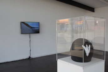 In the foregorund a black crash helmet with a white feather sits in a glass case, facing an LCD monitor showing a steep sand dune
