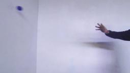 A hand throws a blue ball at a white wall.