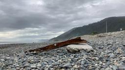 A rusty and warped iron beam rests on coastal rocks. A digger is working in the background.