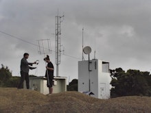 Two people stand by a communication tower holding radio equipment and headphones.