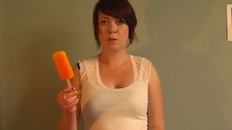 A person holds an orange popsicle up while looking into the camera.