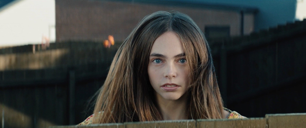 A girl's face appears over a suburban fence, she stares directly at the camera. Her hair appears to rise, as if she has bounced on a trampoline to appear above the fence.