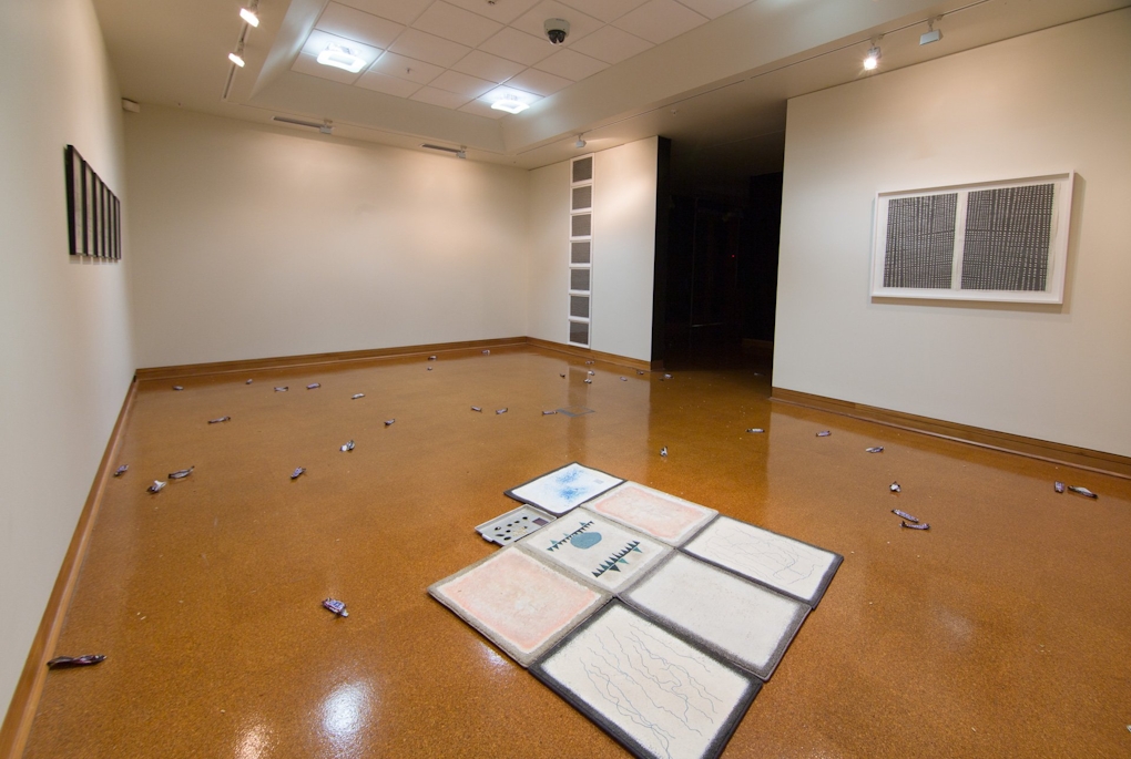 Snickers candy wrappers lie strewn across the gallery floor and around a neat arrangement of open artist sketch books, on the wall are framed geometric drawings