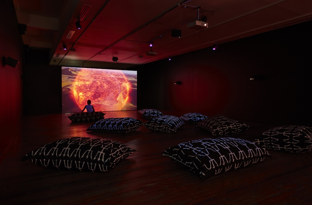 Large patterned cushions are scattered across the gallery floor for audiences to sit on whilst watching a video work of a flaming ball