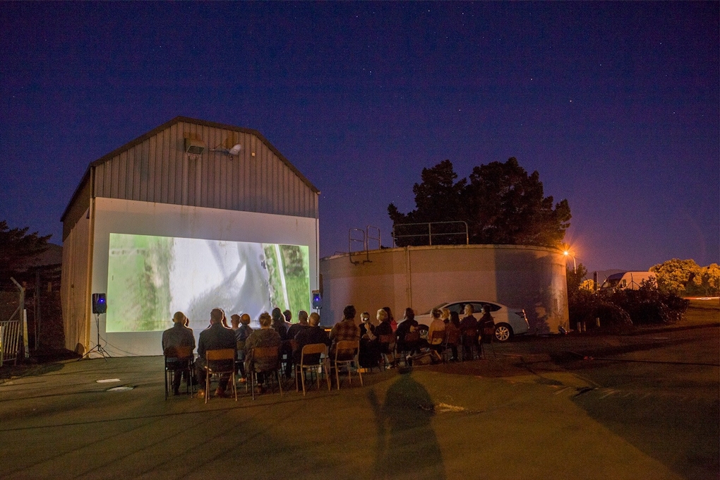An audience watches a film projected on the outside of an industrial building