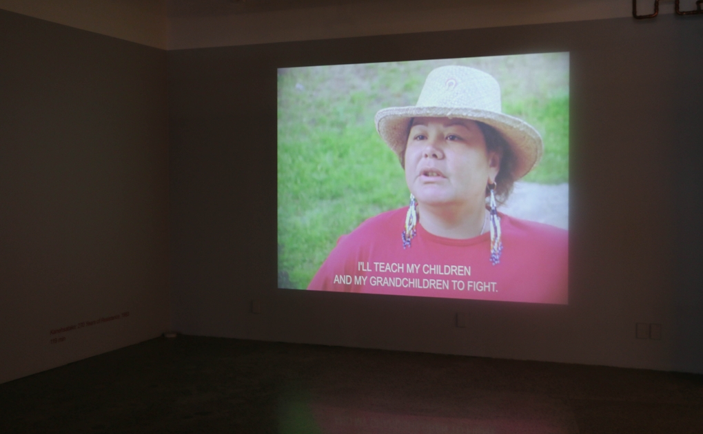 A film is projected onto a gallery wall. The still shows a woman speaking with the subtitle 'I'LL TEACH MY CHILDREN AND MY GRANDCHILDREN TO FIGHT.'