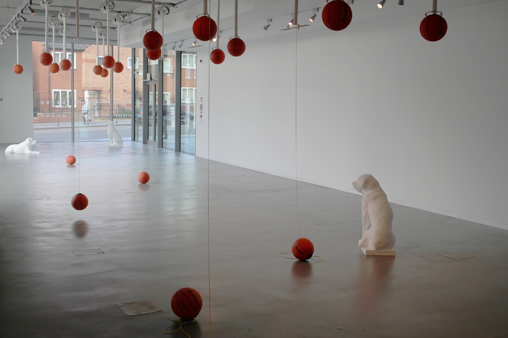 Three white life sized dog sculptures sit on the concrete floor of a gallery. Basketballs are held by contraptions on the ceiling and some of them have fallen to the floor.