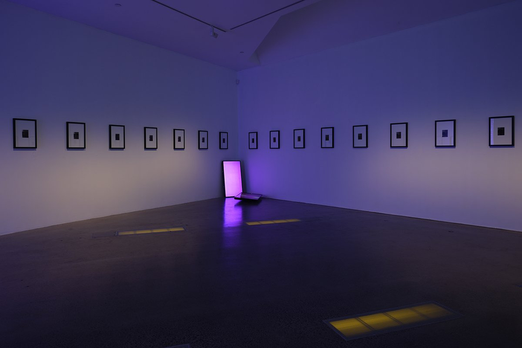 In the corner of a hushed gallery, two LCD screens each show a warm pink colour field, above them a series of framed images fan out from the corner