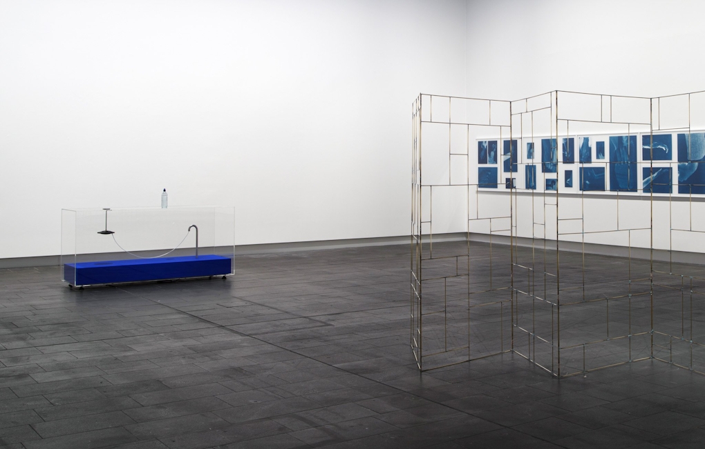 Three artworks in a room, including, at left, a clear cabinet with a bottle of water on top and an electrical transducer hanging inside. At right, in the foreground a free-standing golden room divider. On the wall, a series of blue images showing hands gesturing.