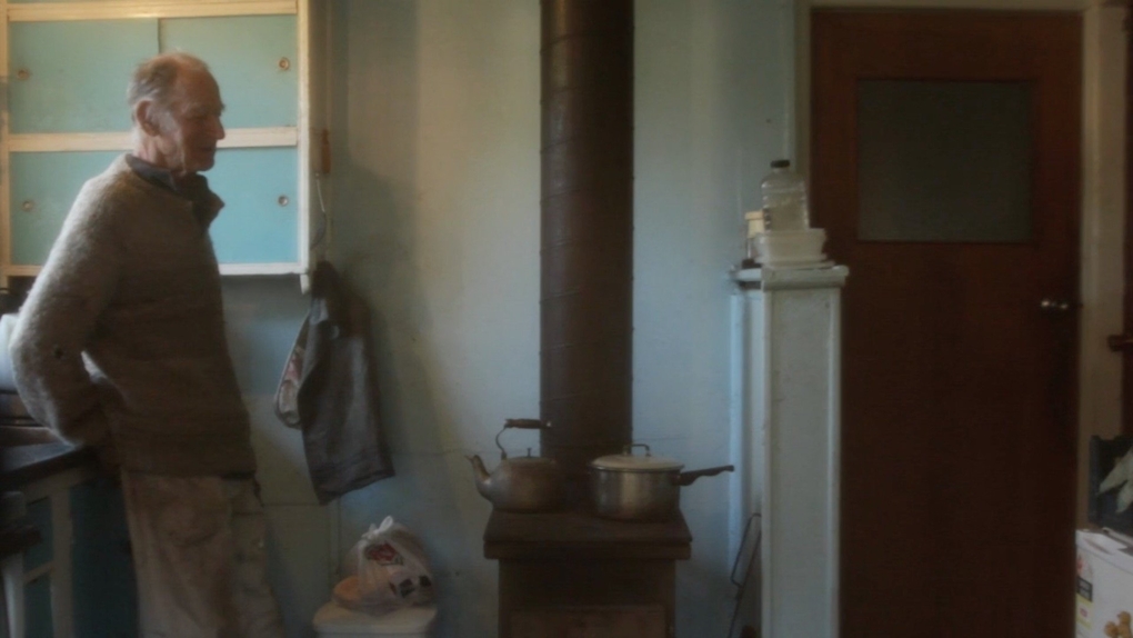 An older man leans against the kitchen counter in a humble house, two pots sit on the wood stove heating up.