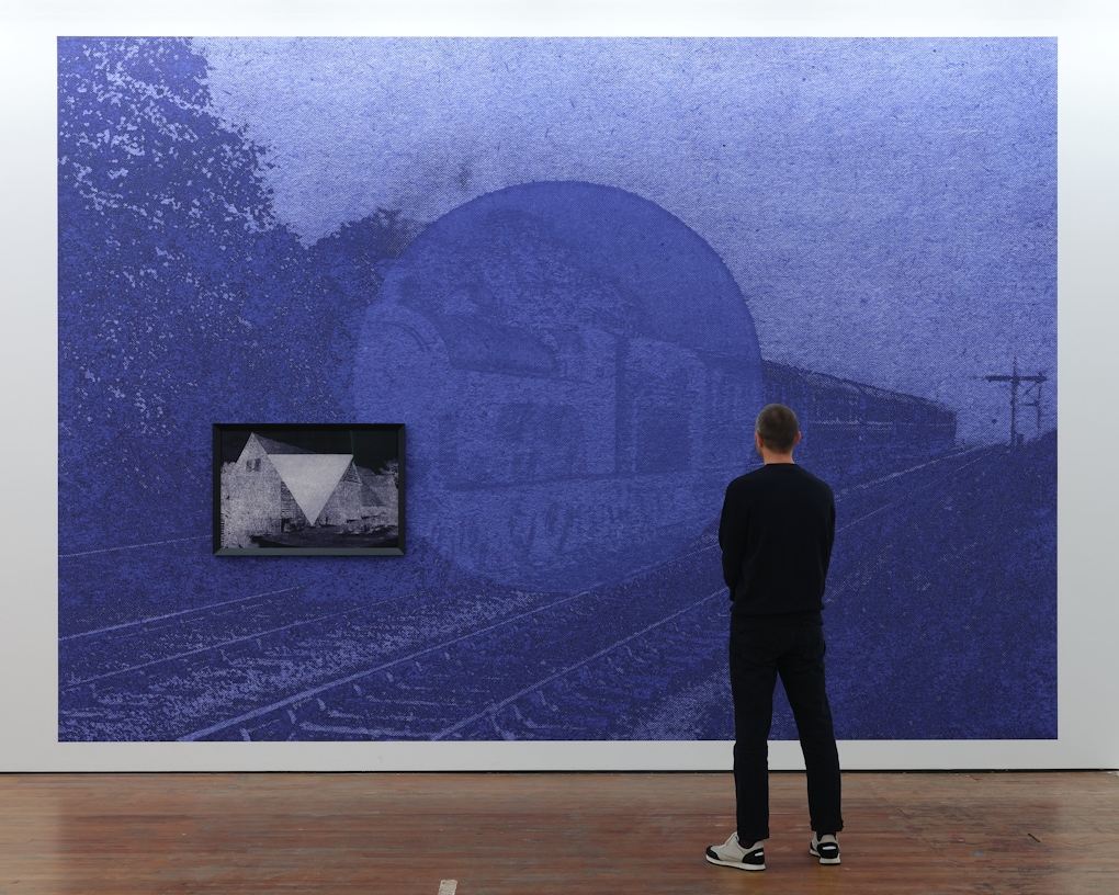 A person stands in front of a blue solarised image of a train on a track, with a large circle obscuring the centre of the frame as if suggesting another presence