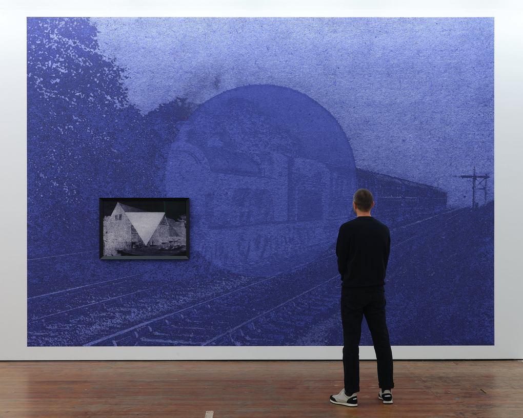 A person stands in front of a blue solarised image of a train on a track, with a large circle obscuring the centre of the frame as if suggesting another presence