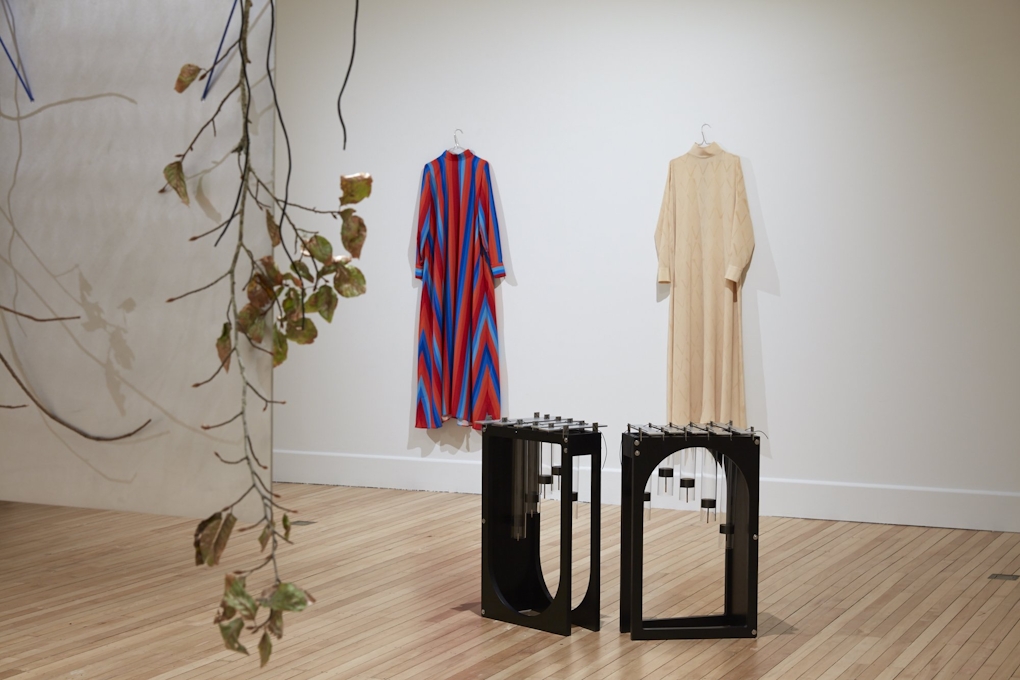 Two dresses hang on the wall, each modest but possibly ceremonial, in the foreground is a hanging branch with leaves, in the mid ground are two percussion instruments