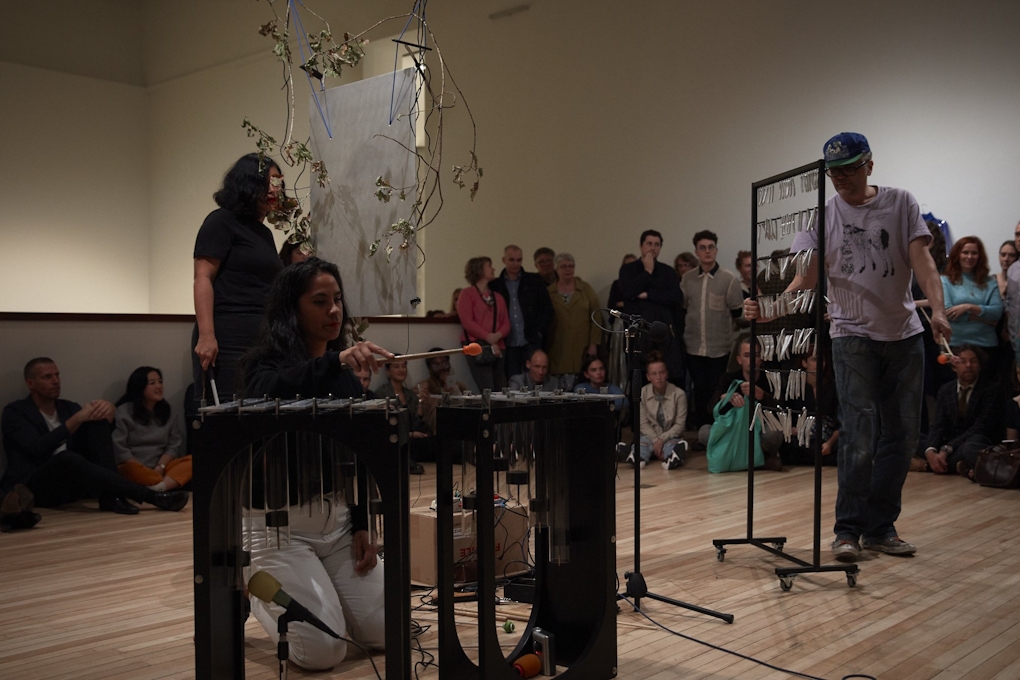 Three musicians play percussion instruments in a gallery, watched by onlookers