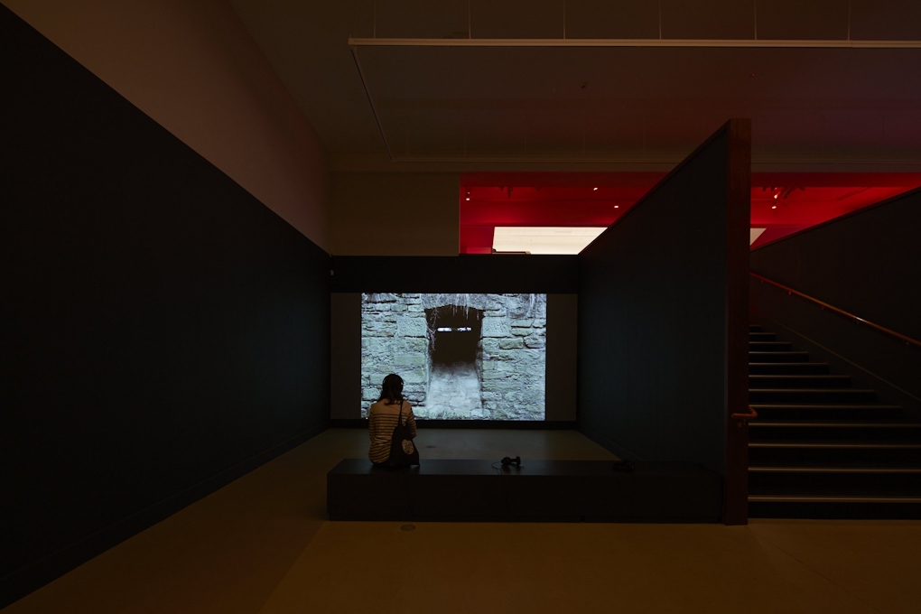 Someone sits watching a projected film showing a dark entrance to a stone structure
