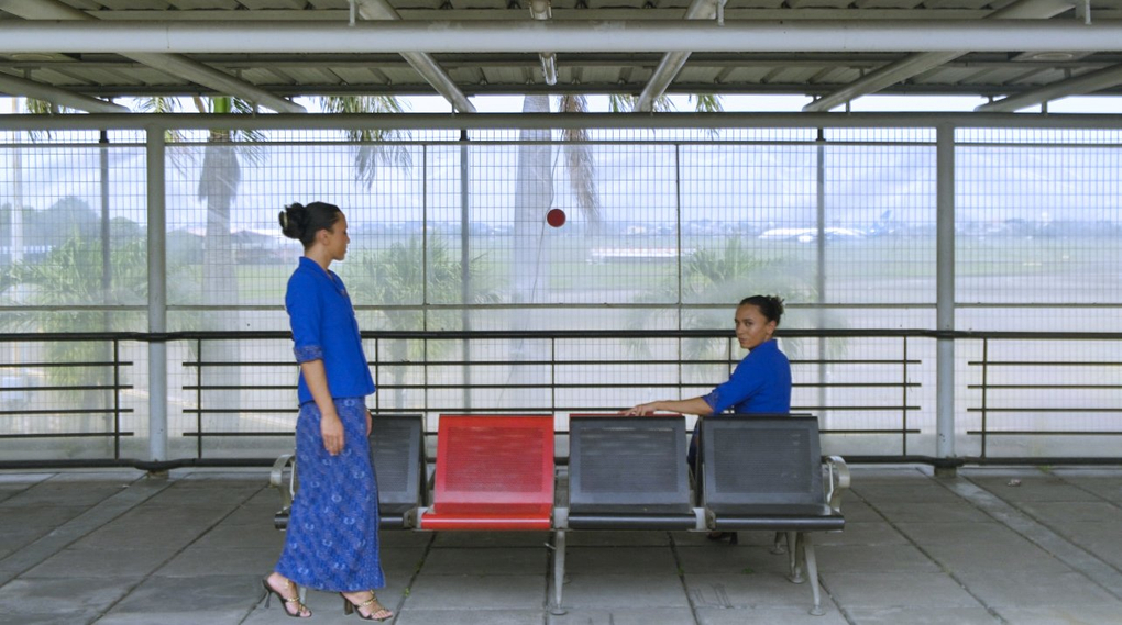 Two women (one standing, one seated) are wearing traditional Indonesian dress in an open-air airport waiting area