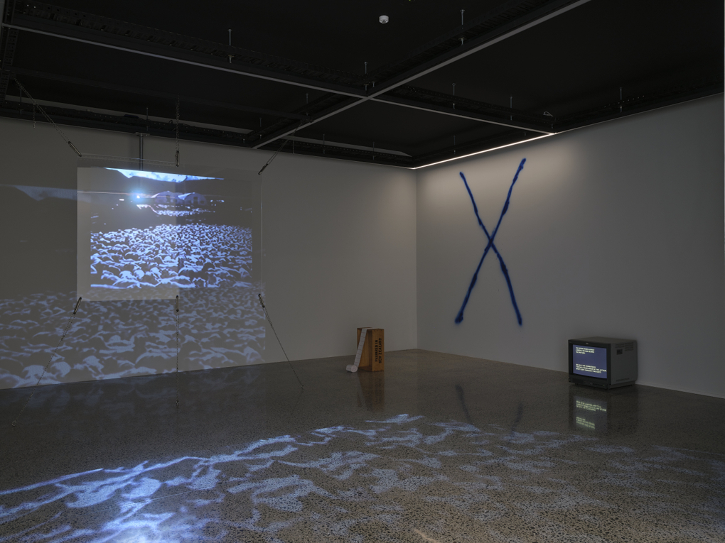 A gallery showing four distinct artworks in an installation. At left we see a projection screen displaying an image of a mob of sheep herded tightly together. The image spills through the screen and onto the wall where it is reflected onto the floor. The second artwork is a wooden box sat on it's end with a roll of paper cascading upwards to the top. Thirdly, a large blue X has been spraypainted on the wall. The 4th and final work is a CRT video monitor which sits on the floor and shows yellow text.