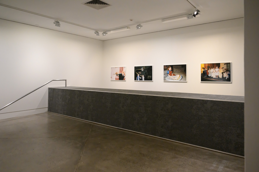 4 pastel drawings hang on a wall; in front is a grey carpeted divider that bisects the exhibition space