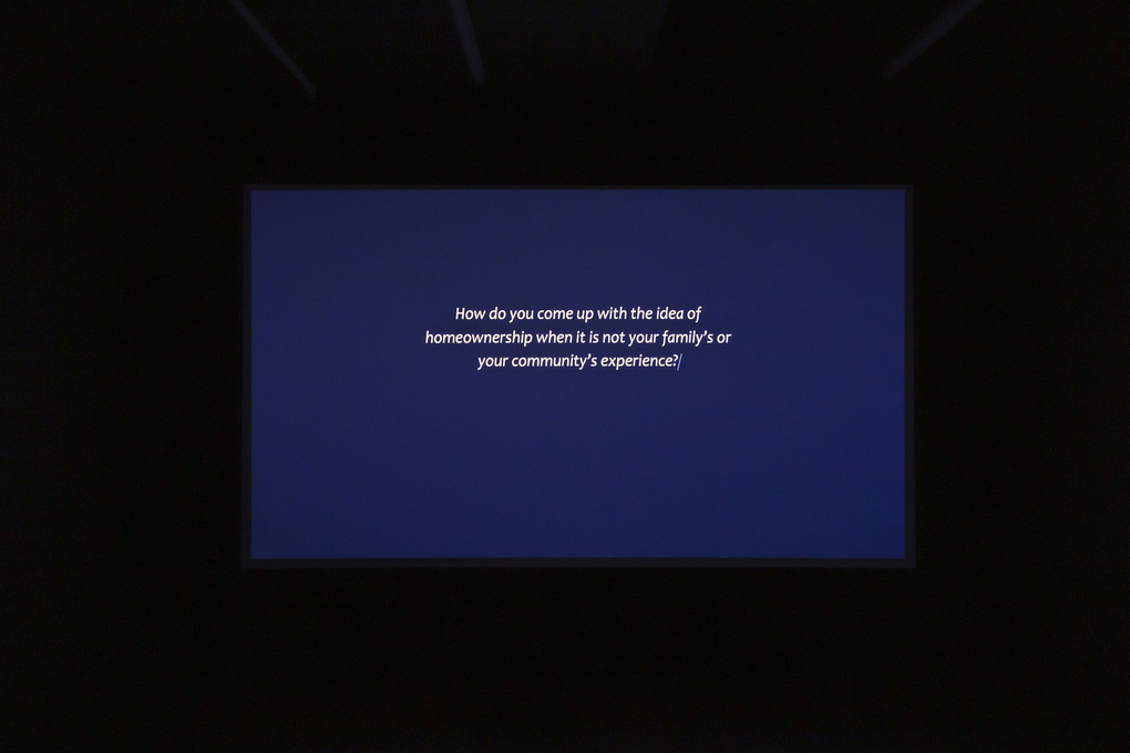 A blue screen in a darkened gallery room, with text onscreen: "How do you come up with the idea of home ownership when it is not your family's or your community's experience?"