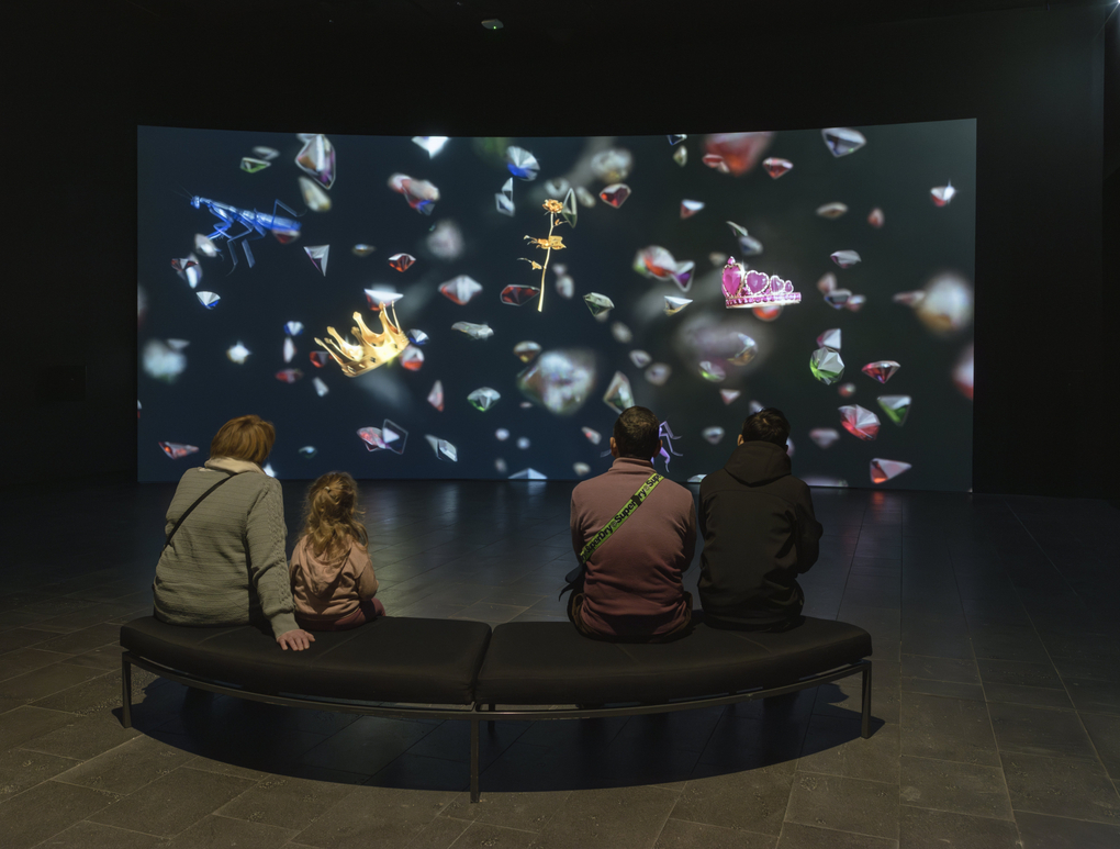 In a dark gallery four people watch an animated projection of objects floating in black space, including a golden crown, a pink tiara and jewels.
