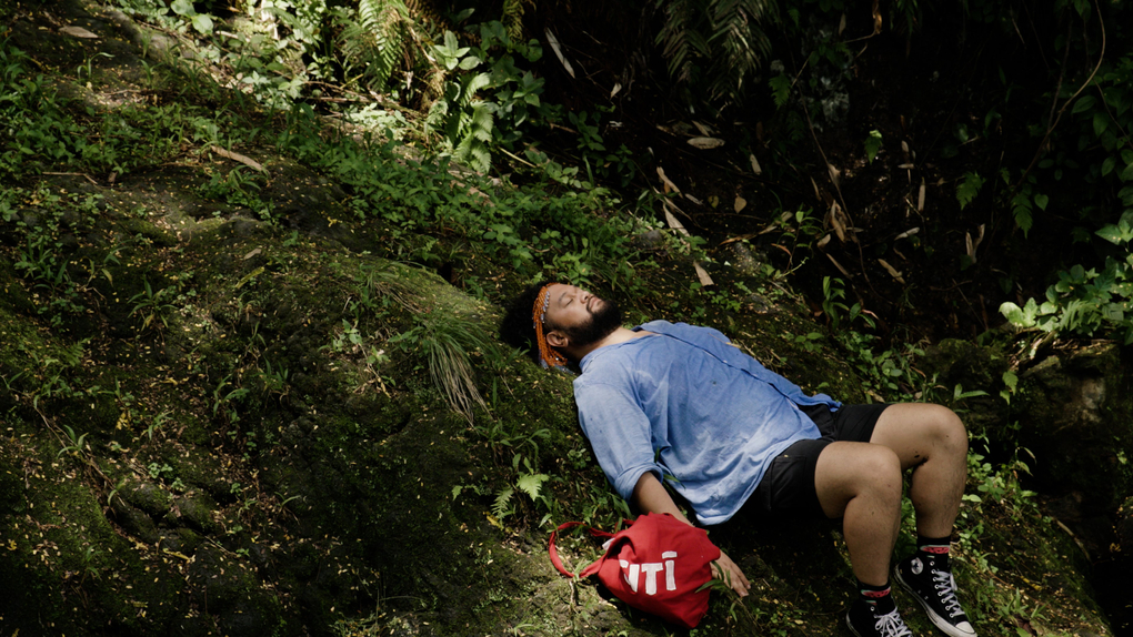 The artist lies on a mound in dense bush, wearing a signature beaded headdress