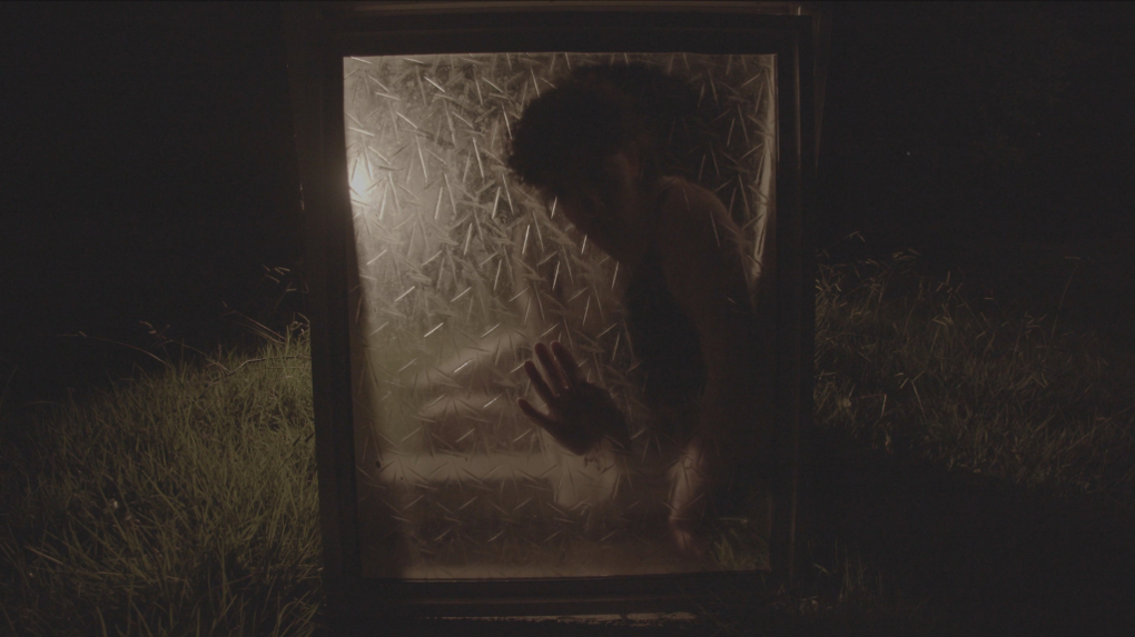 A frosted window is propped up in a bed of grass. Behind it is a silhouette of someone touching the window.