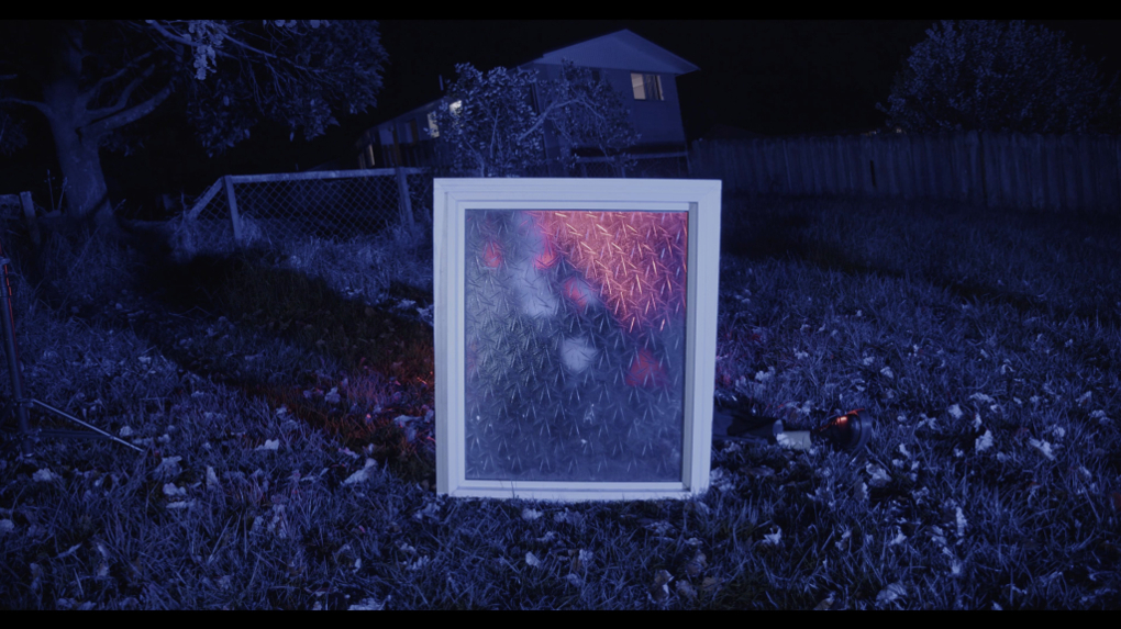 A frosted window is propped up in a backyard lit up in blue. Behind the window is the blurred outline of a person. Behind them is a red light.