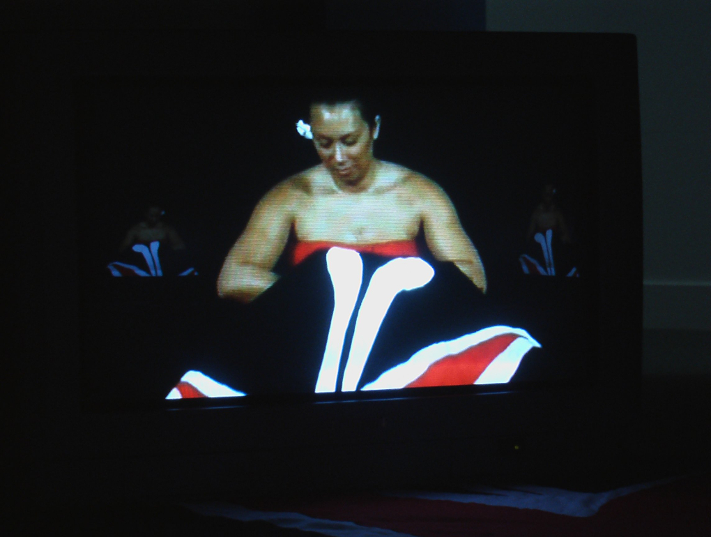 An image of a video of a woman looking down at the tivaevae she is sewing. She has a pareu wrapped around her and wears a flower behind her ear.