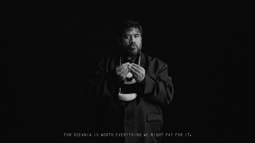 An Pasifka interpreter faces the camera and uses their hands and expressions to convey their message in sign language. Text at the bottom of the screen says "FOR OCEANIA IS WORTH EVERYTHING WE MIGHT PAY FOR IT".
