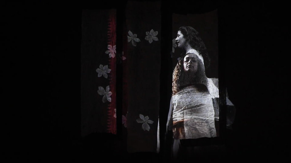 Three panels of barkcloth display a projection of two overlapping female figures. One figure faces forward while the other is shown in profile, surrounded by pale floral motifs and a serrated red rectangle against a black background.