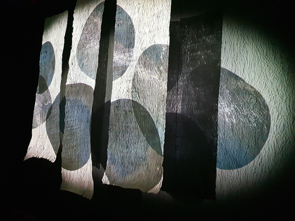 Projected onto three panels of barkcloth are various circular grey shapes. The texture of the barkcloth catches the projection and spreads across the dark installation space.