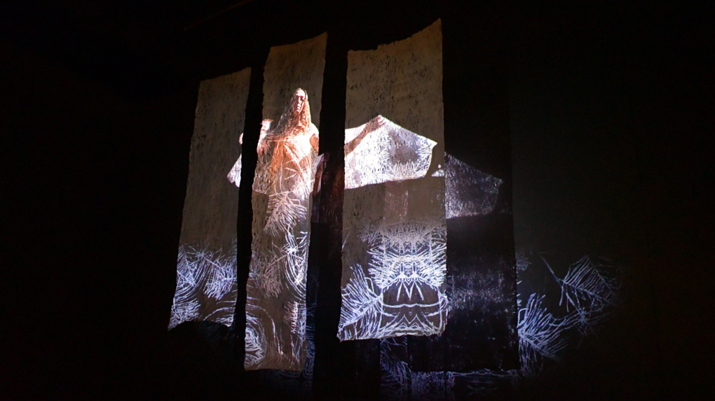 Three panels of barkcloth display a projection of a standing figure with arms outstretched, overlaid with white linework that spreads across the dark installation space.