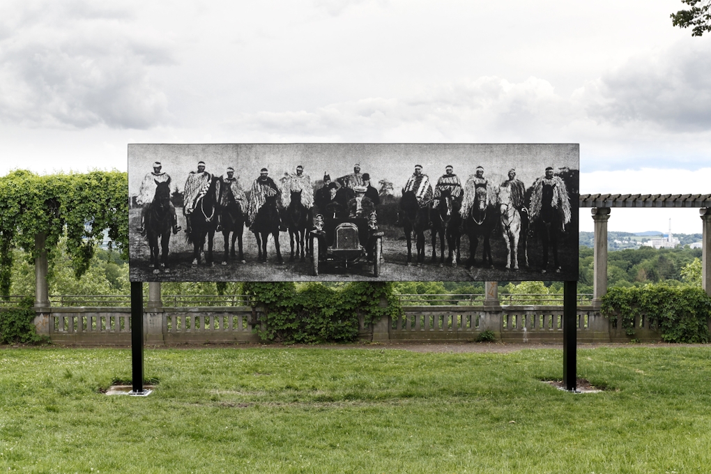 A billboard in a park with an old photo of people wearing korowai on horseback