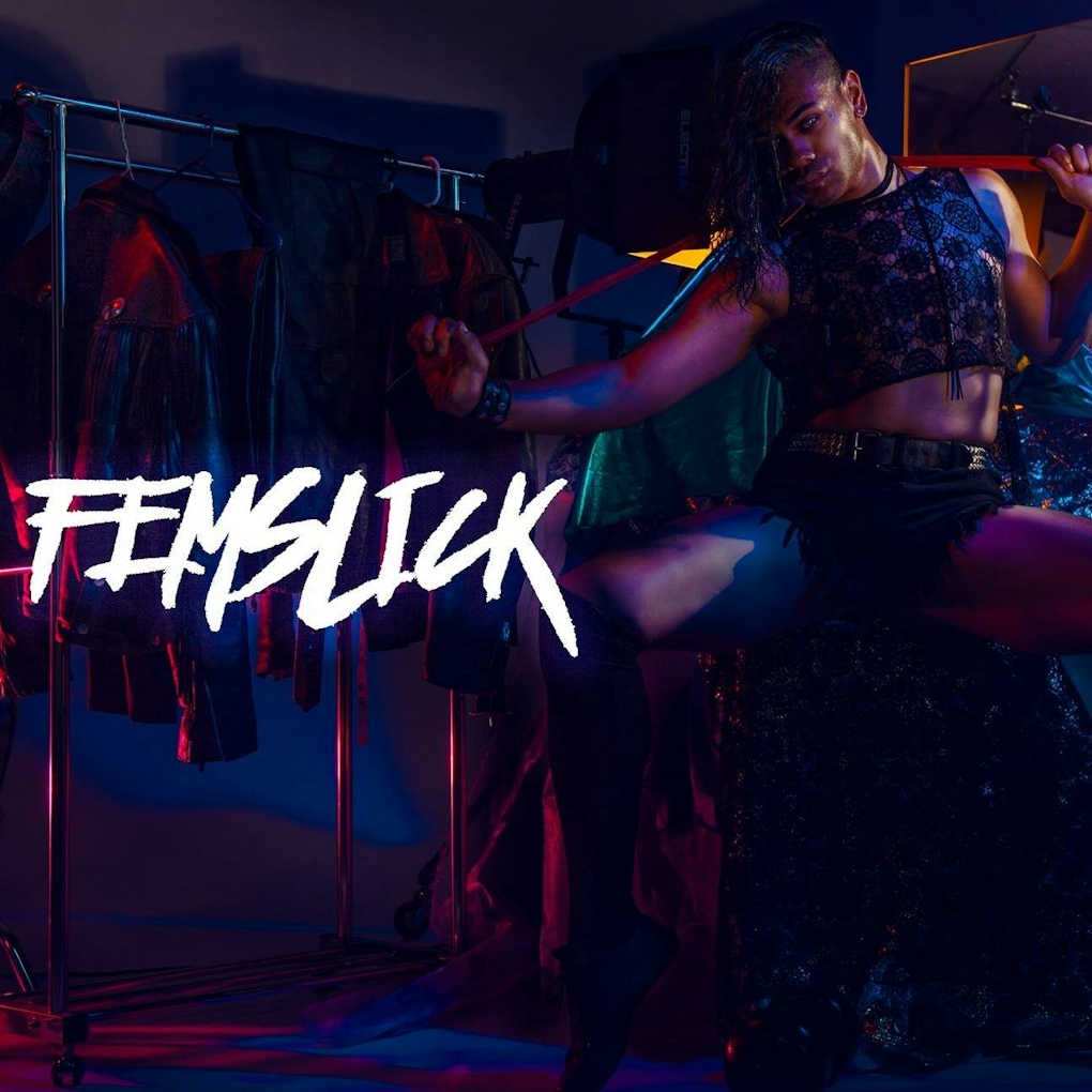A dramatically lit person sits on a stool with their legs spread apart holding a ribbon with out stretched hands. The words FEMSLICK are written ontop of the image.