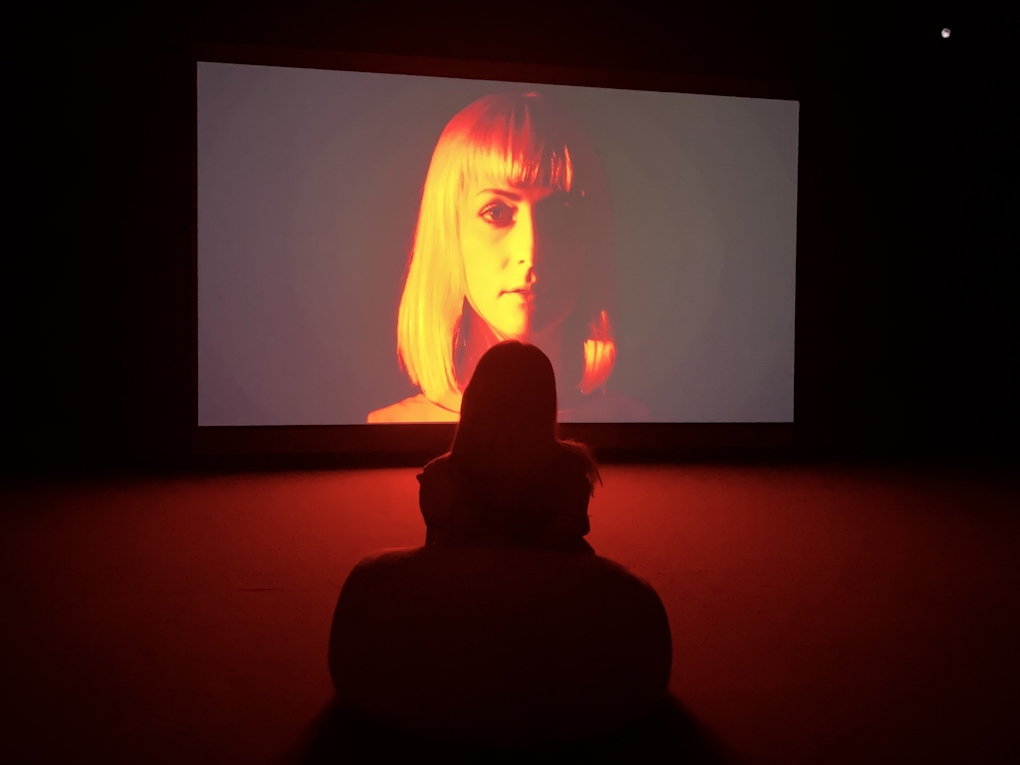 Installation view of Marianna Simnett exhibition, showing a viewer in the gallery and a shadowy woman's face with red hue on screen