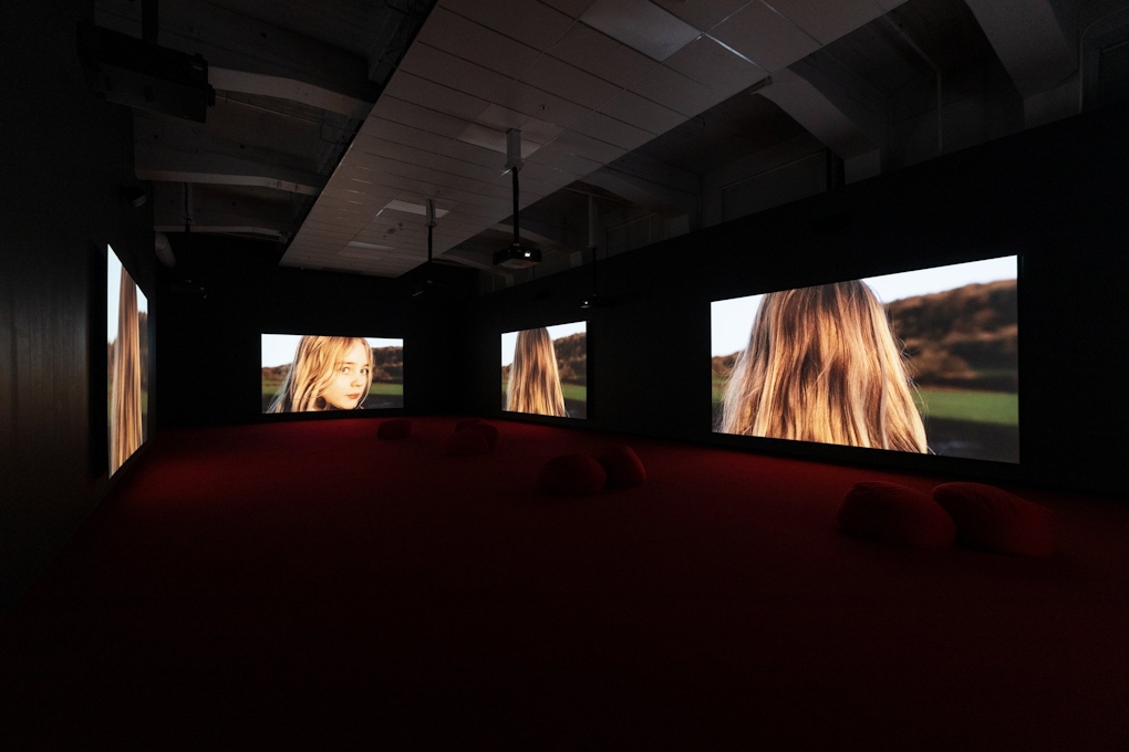 Four large screens in a room show the scene from different angles, at the same time. A young girl walks along a field, showing the back of her head, the side of her face, the back of her head.