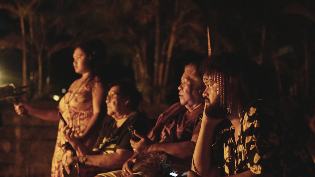 The artists and their family watch the performer spin a nifo oti (fire knife) at night with recording equipment