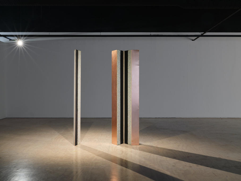 Three tall objects resembling thick, copper clad planks stand on their ends in a gallery. At right two are placed as if hinged. The inside of each appears to be foam.