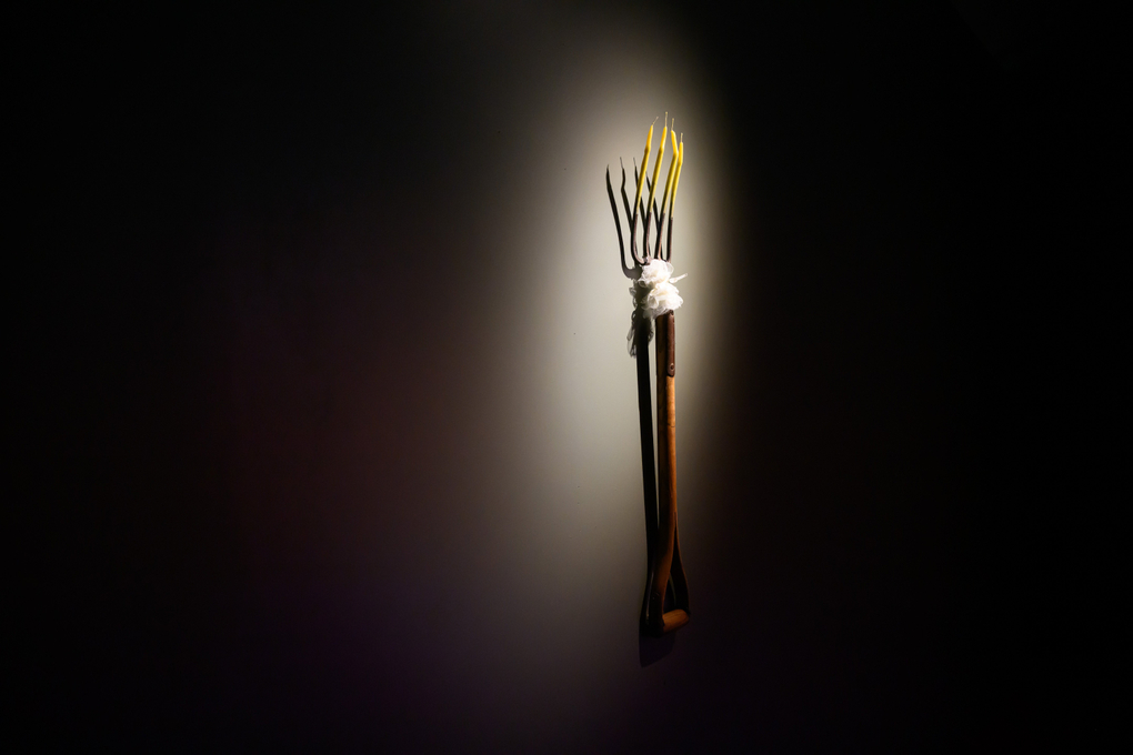 A makeshift pitchfork with candles for prongs hangs on a gallery wall