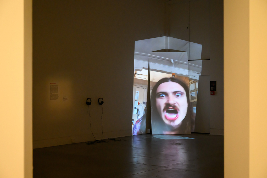 A person sings open-mouthed to camera, projected onto a corner wall that distorts the shape of the image like a broken mirror
