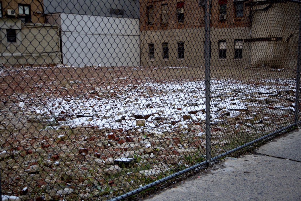 An area of rubble, behind a metal fence, covered in a white substance which could be snow. A brick building is in the background.