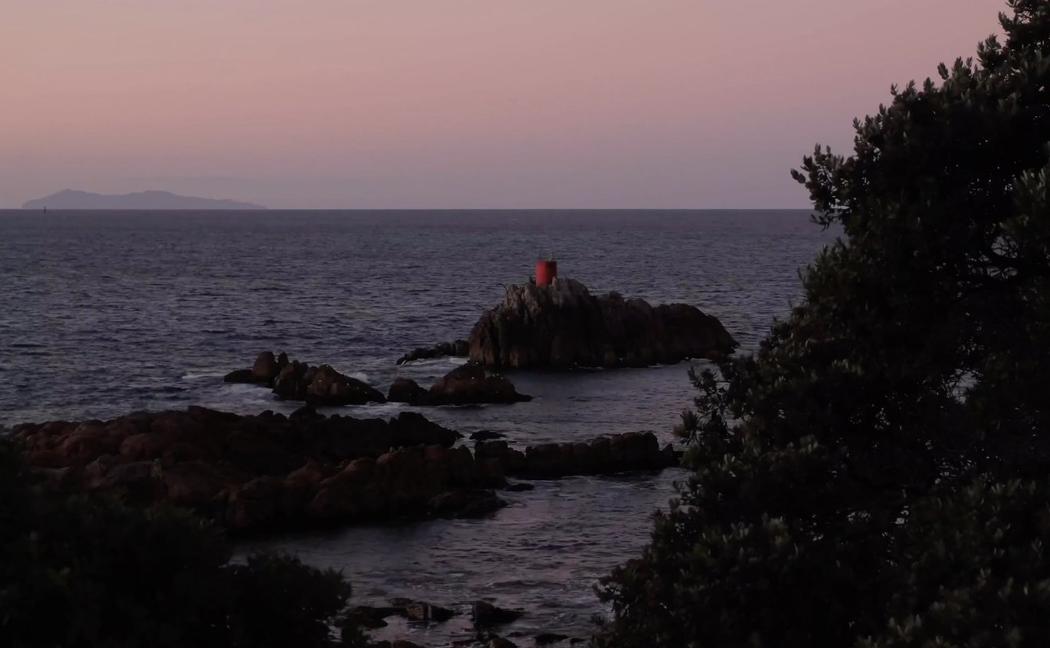 Jagged rocks stick out of the ocean underneath a pinky blue sunset or sunrise. On the rocks is a bright red cylinder object.