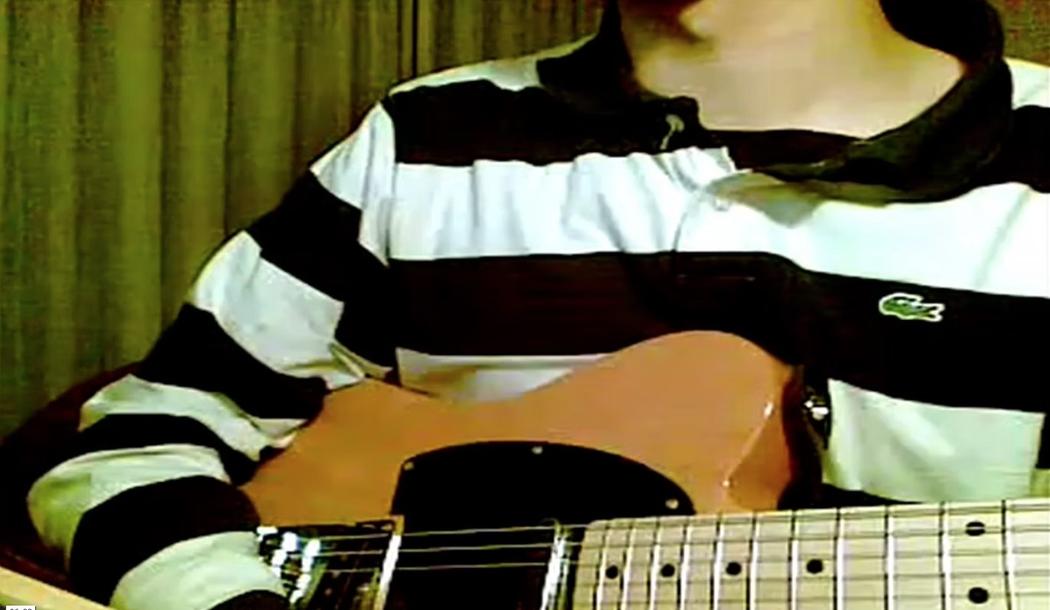 A pixelated image of a person holding a guitar wearing a long sleeve polo shirt, behind them are green curtains.