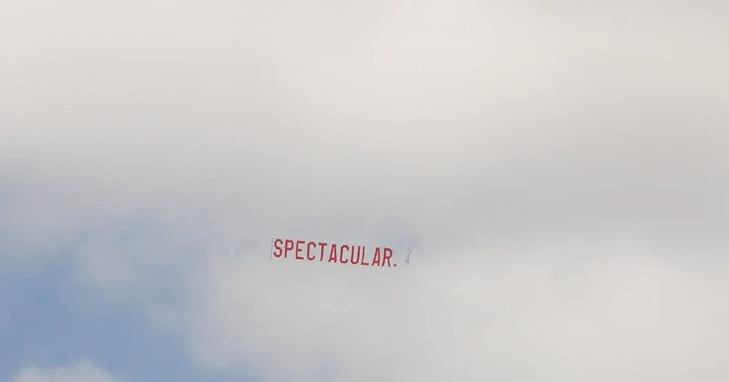 A large red ariel banner is flown through the air, towed from an out of the shot aeroplane. The text on the banner reads "SPECTACULAR." in large red lettering against a cloudy blue sky