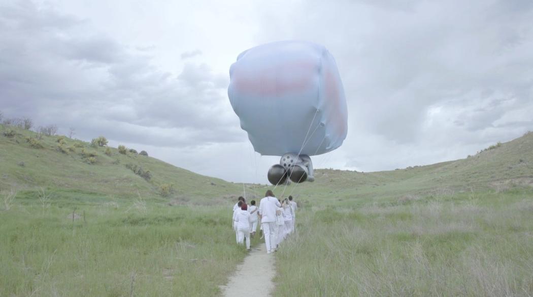 A group of people in white outfits carry the strings of a giant balloon through a grassy landscape