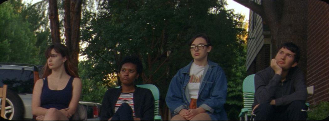 Four teenagers sit on the footpath in a row in deckchairs, gazing out into the distance.