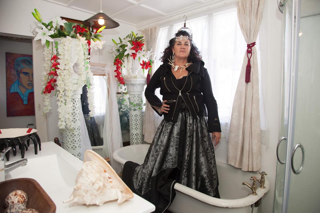 Suzanne stands up in her bathtub wearing a silky long dress, black jacket and traditional necklace and headpiece. She presents as powerful, serious, cool and confident